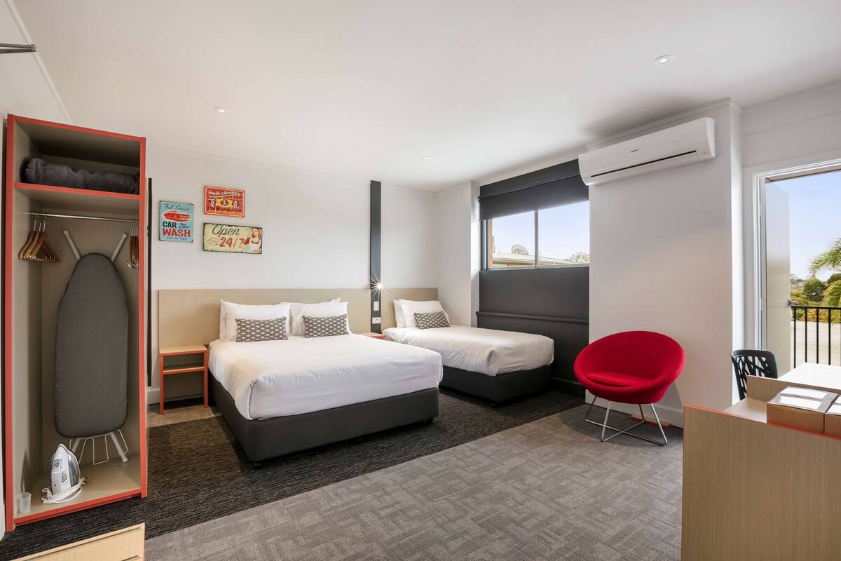 A modern hotel room with two beds, a red chair, a desk, and an ironing board.