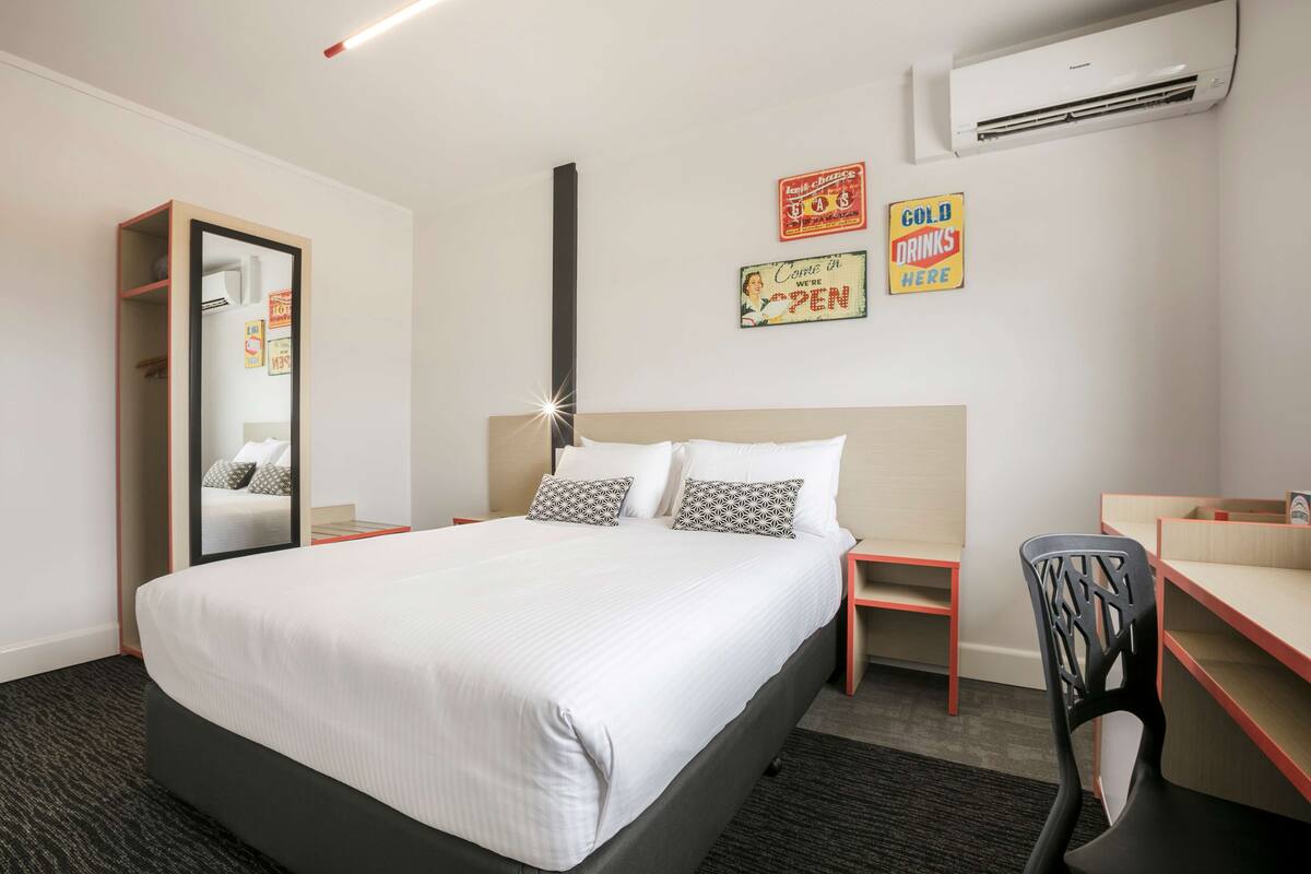 A hotel room with a large bed, a desk, a chair, a wardrobe, and an air conditioning unit.