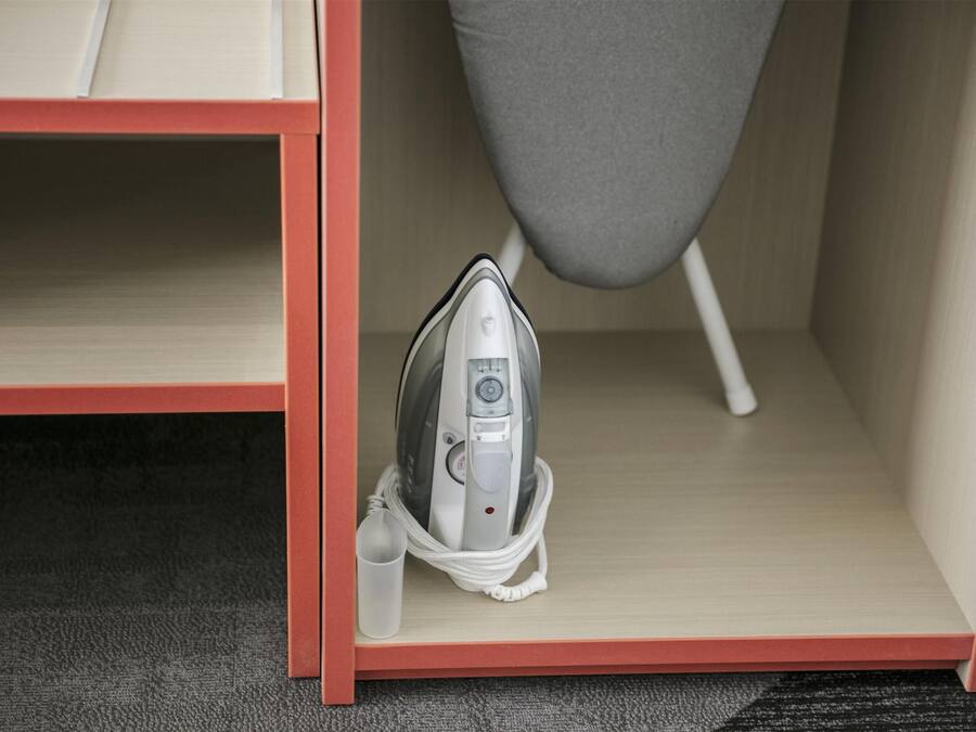 Una habitación con una estantería de marco rojo, una silla gris y un planchado en una bolsa.
