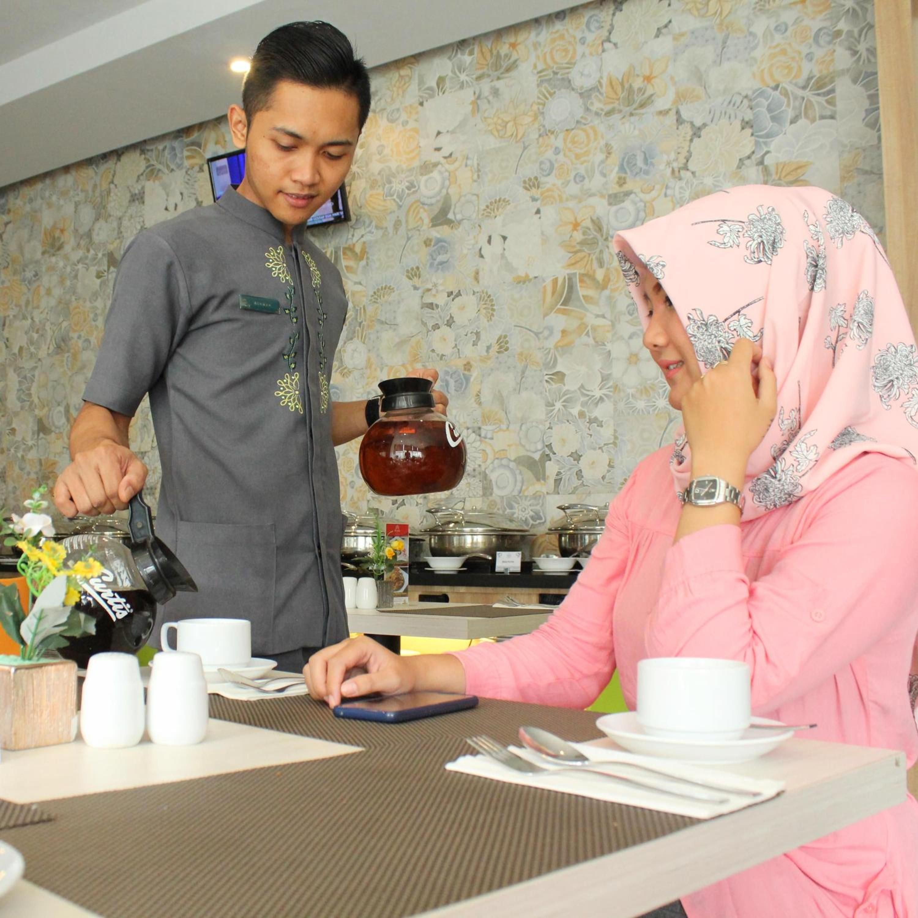 A person serving tea to another person at a table with a floral wallpaper background.