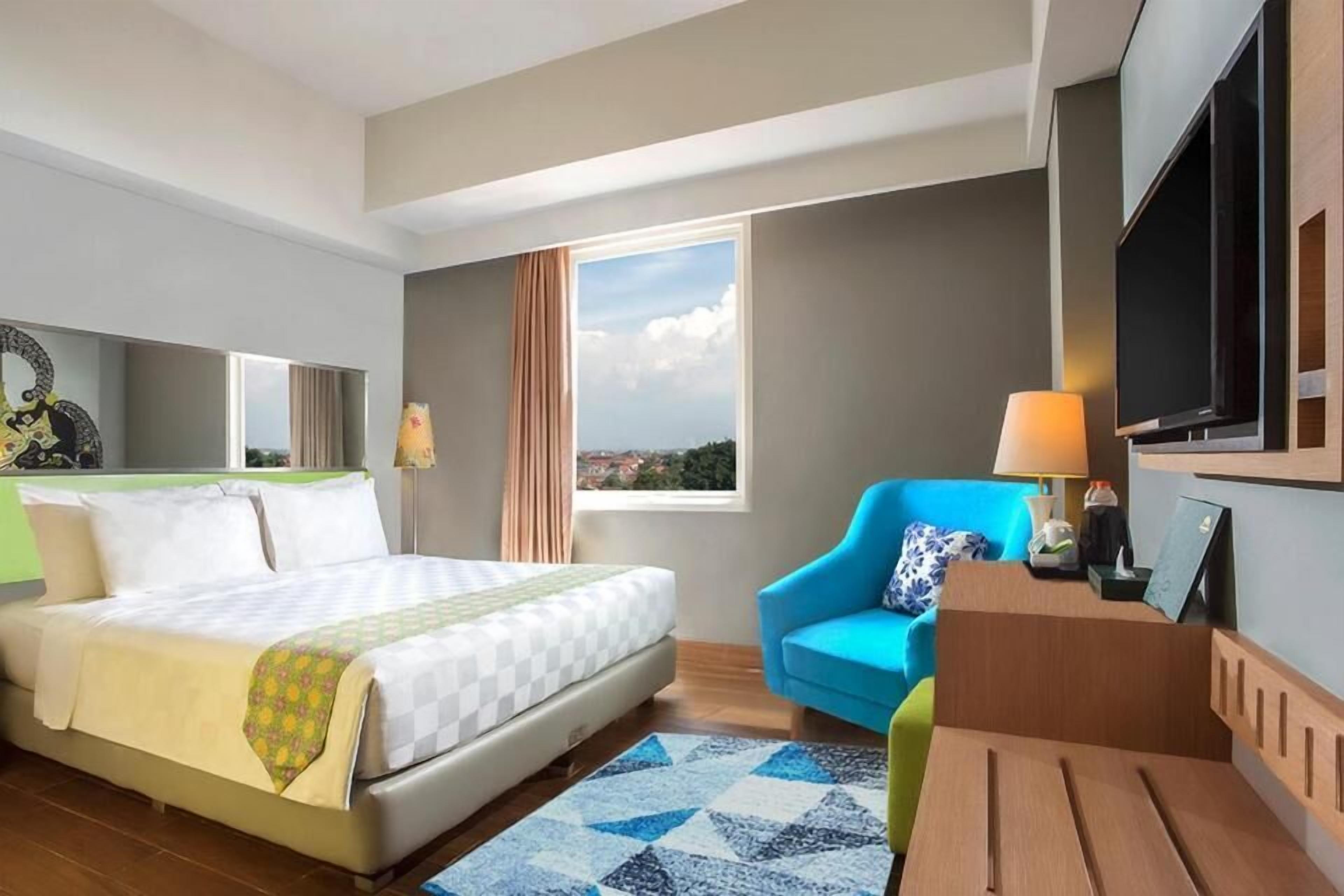 Une chambre d’hôtel avec un lit, un fauteuil bleu, un bureau, une télévision et une fenêtre donnant sur un beau paysage.