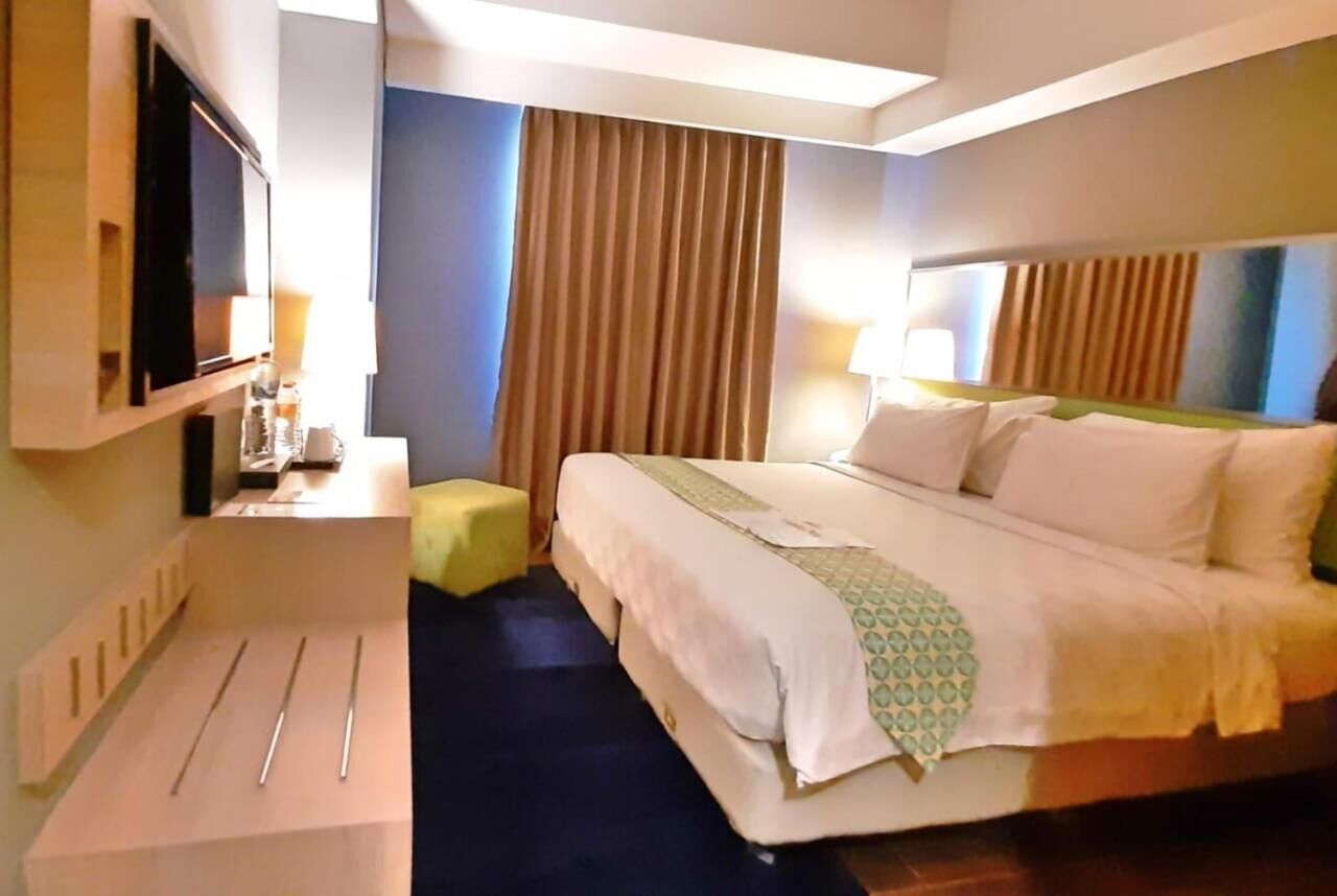 A hotel room with a bed, a TV mounted on the wall, a desk with a lamp, and a window with curtains.