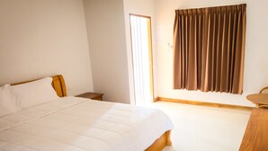 A bedroom with a large bed, a wooden nightstand, a small table, and a window with curtains. - Wangthatum (Si Maha Phot)