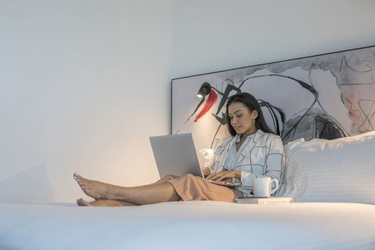 A person using a laptop on a bed with a cup, in a room with a large abstract painting on the wall.