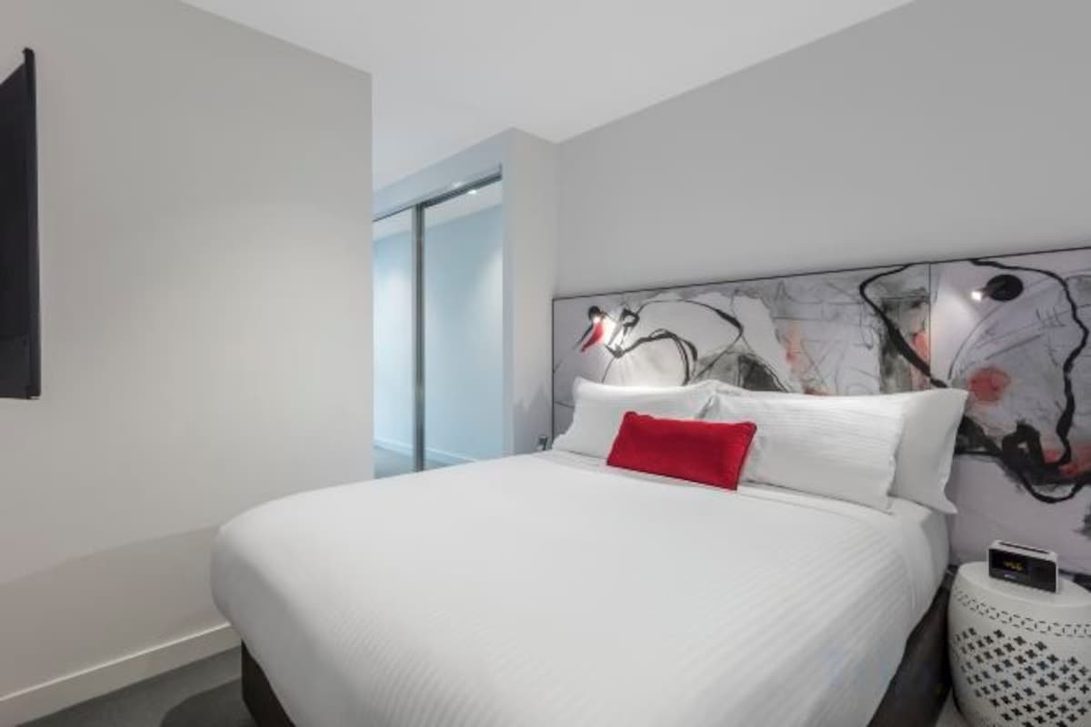 Una habitación de hotel moderna con una cama grande, un cabecero con arte abstracto, una almohada roja y una mesita de noche con lámpara.