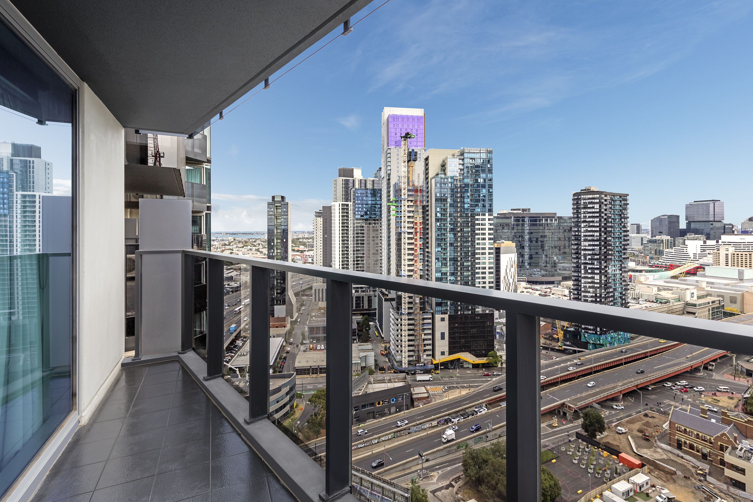 A cityscape view from a balcony with modern buildings, a highway, and a clear sky.