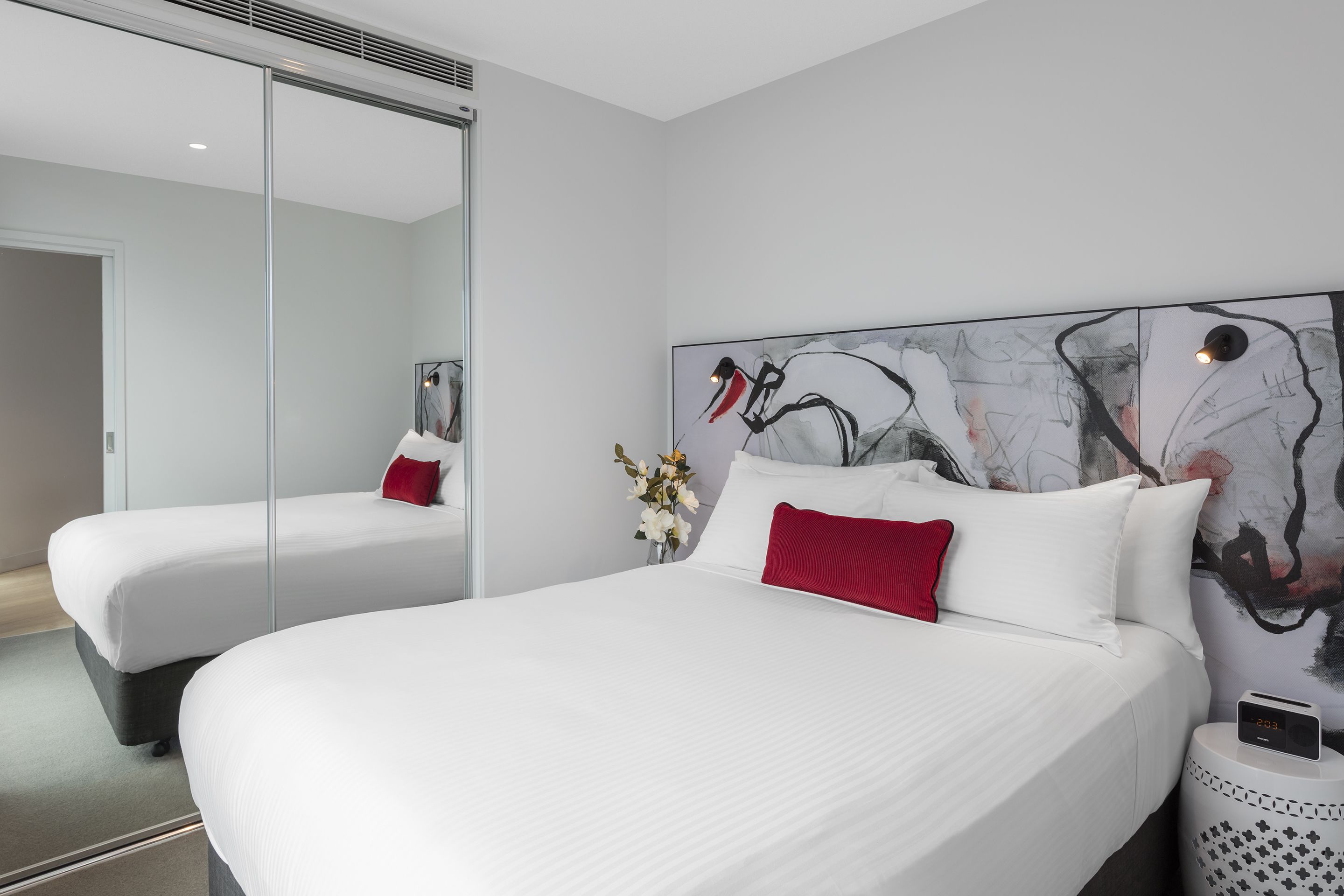 Un hotel moderno con dos camas, un espejo grande, arte abstracto en la pared, un jarrón con flores y un reloj digital en una mesita de noche.