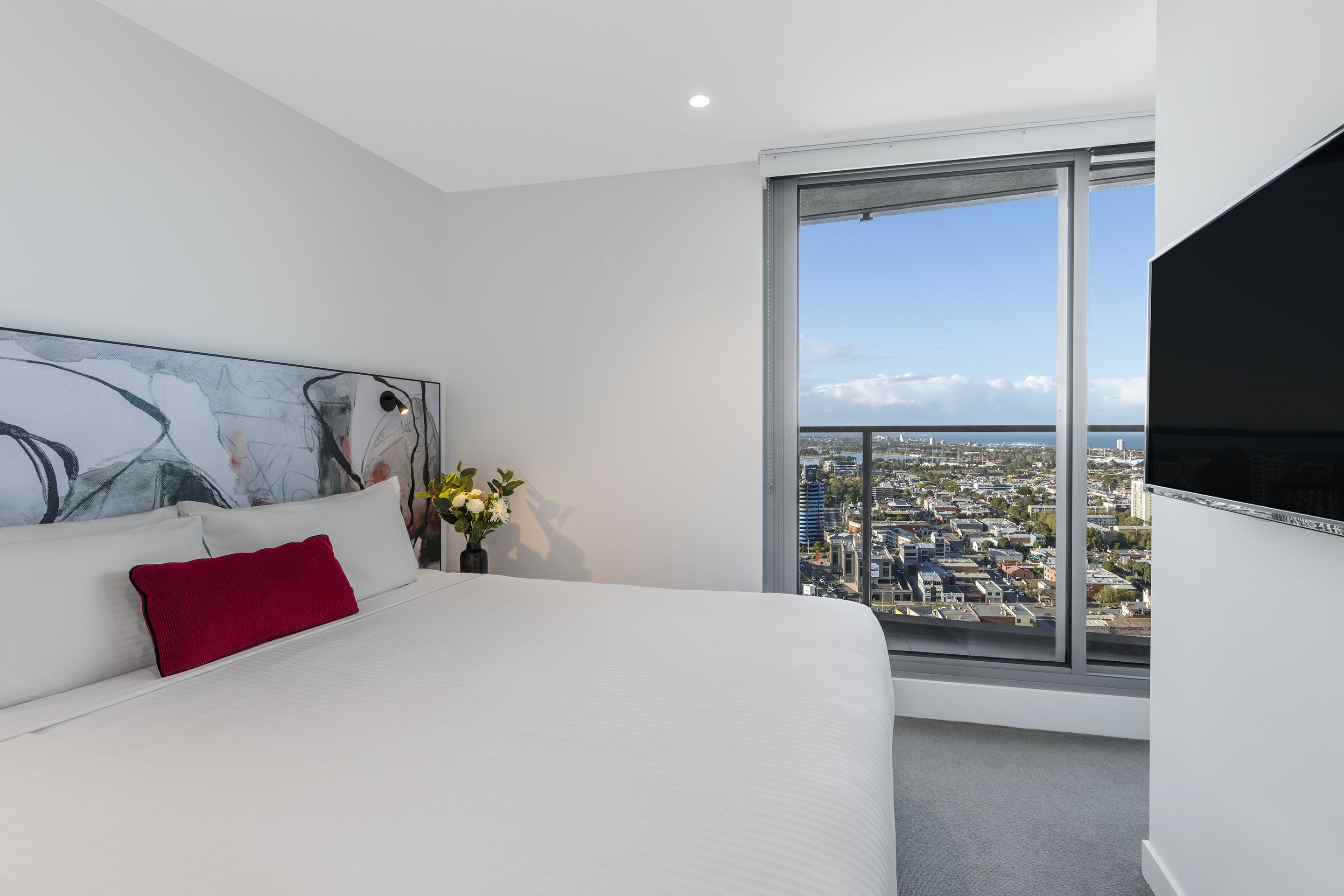 Une chambre d’hôtel moderne avec un grand lit, un oreiller rouge, un tableau au mur, un vase de fleurs et une vue sur la ville par une grande fenêtre.