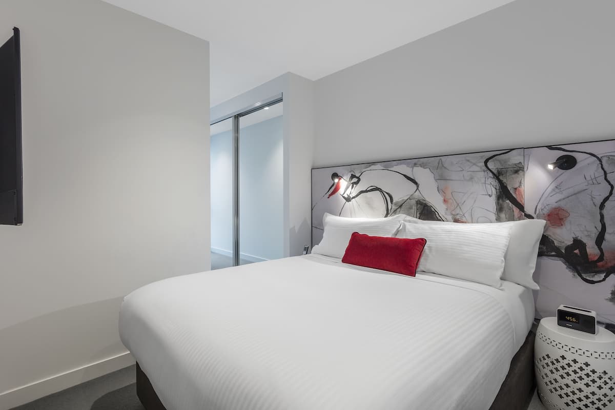 Una habitación de hotel moderna con una cama grande, una almohada roja, un reloj montado en la pared y arte abstracto en la pared.