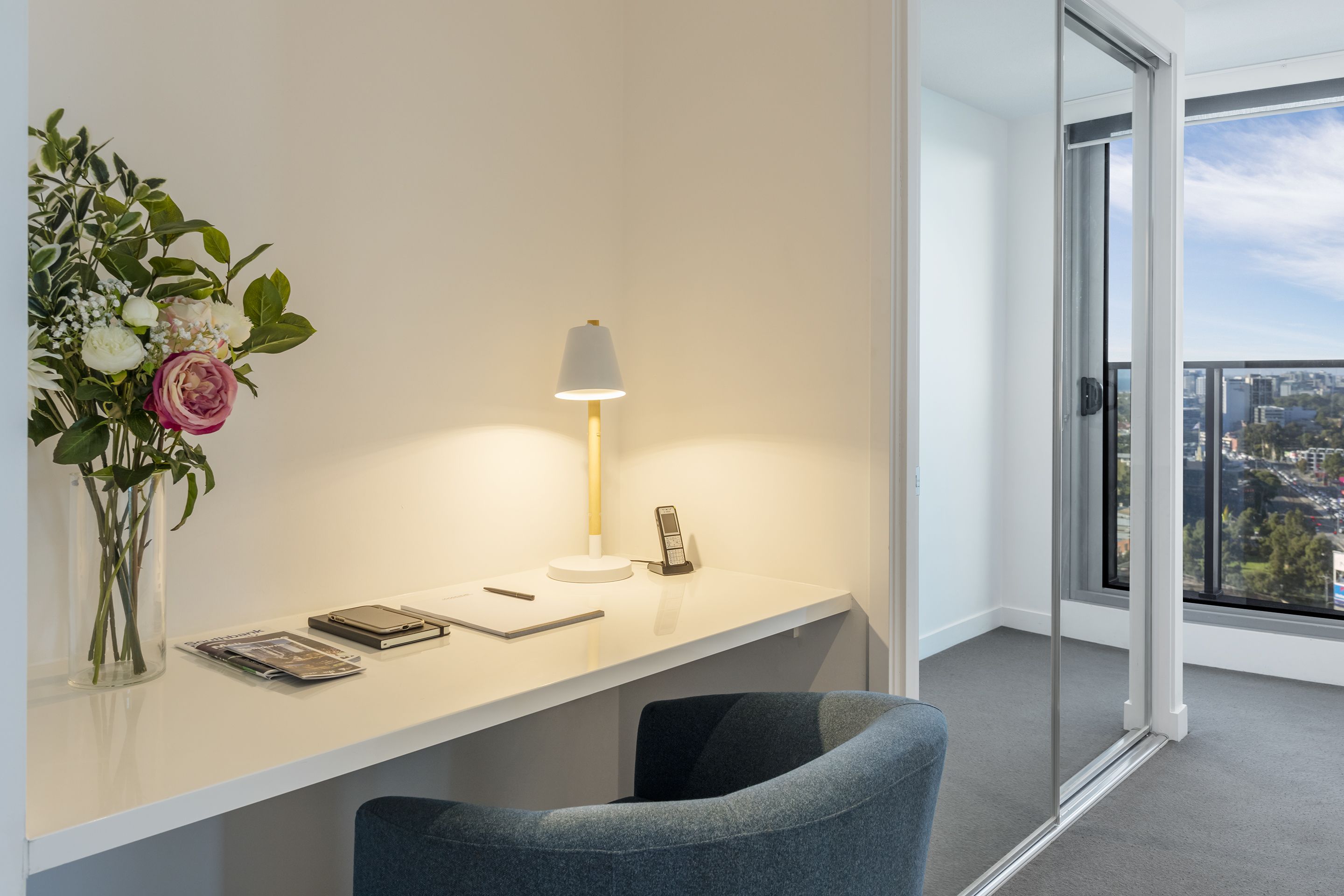 Una habitación de hotel moderna con un escritorio blanco, un jarrón de flores, una lámpara y vistas a la ciudad a través de amplios ventanales.