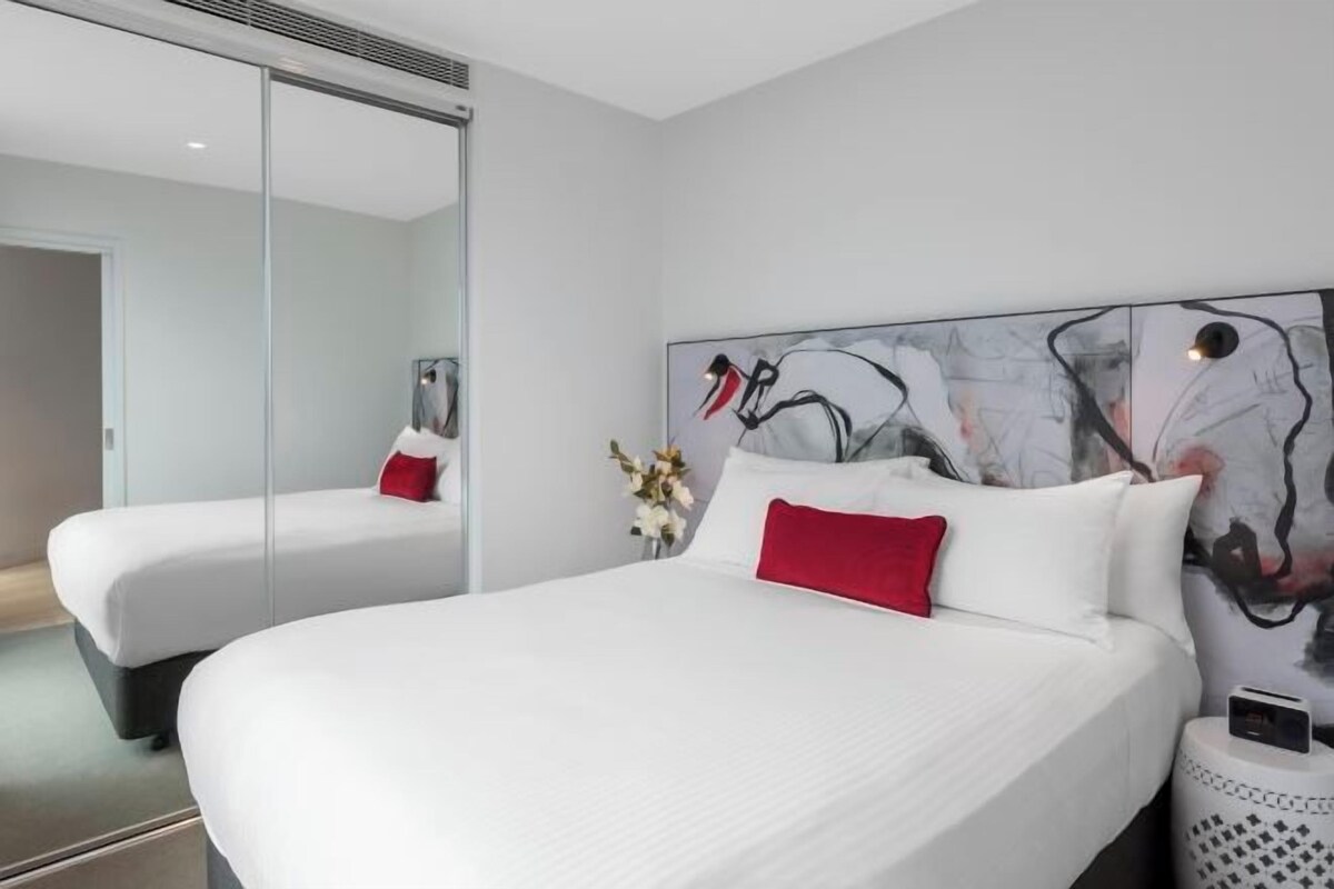 A modern hotel room with a large bed, a bedside table, a mirror, and abstract wall art.