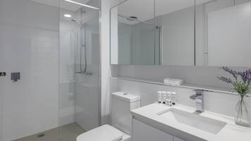 Une salle de bain moderne avec une douche à parois de verre, des toilettes blanches, un lavabo avec un miroir au-dessus et un vase avec des fleurs posé sur le comptoir.