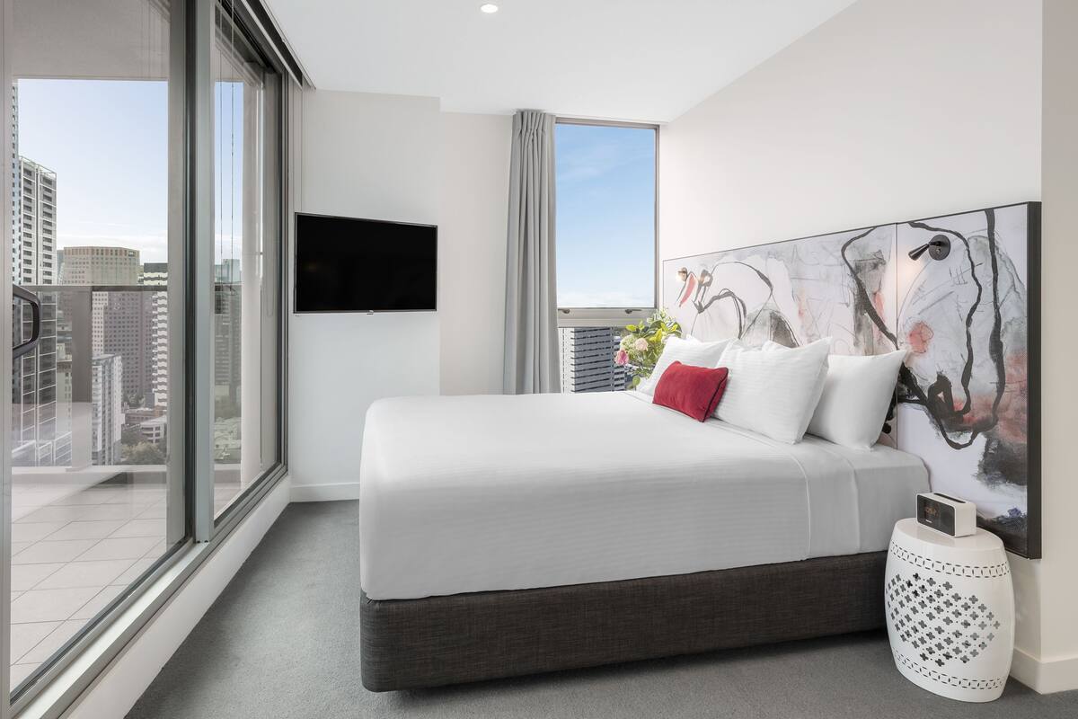 Una habitación de hotel moderna con una cama grande, un televisor de pantalla plana, vistas a la ciudad y un cuadro abstracto de gran tamaño en la pared.