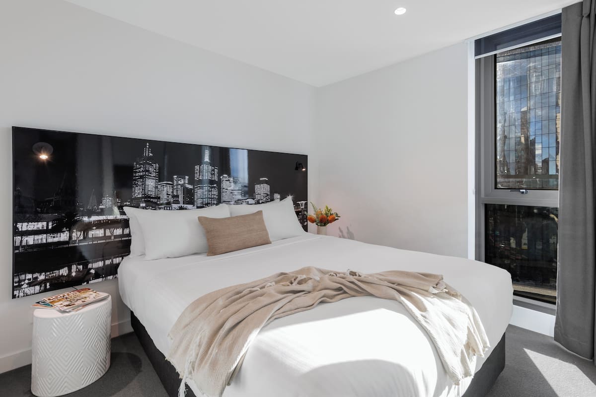 Una habitación de hotel moderna con una cama grande, una obra de arte con un paisaje urbano en la pared, una mesita auxiliar pequeña y una ventana con cortinas.