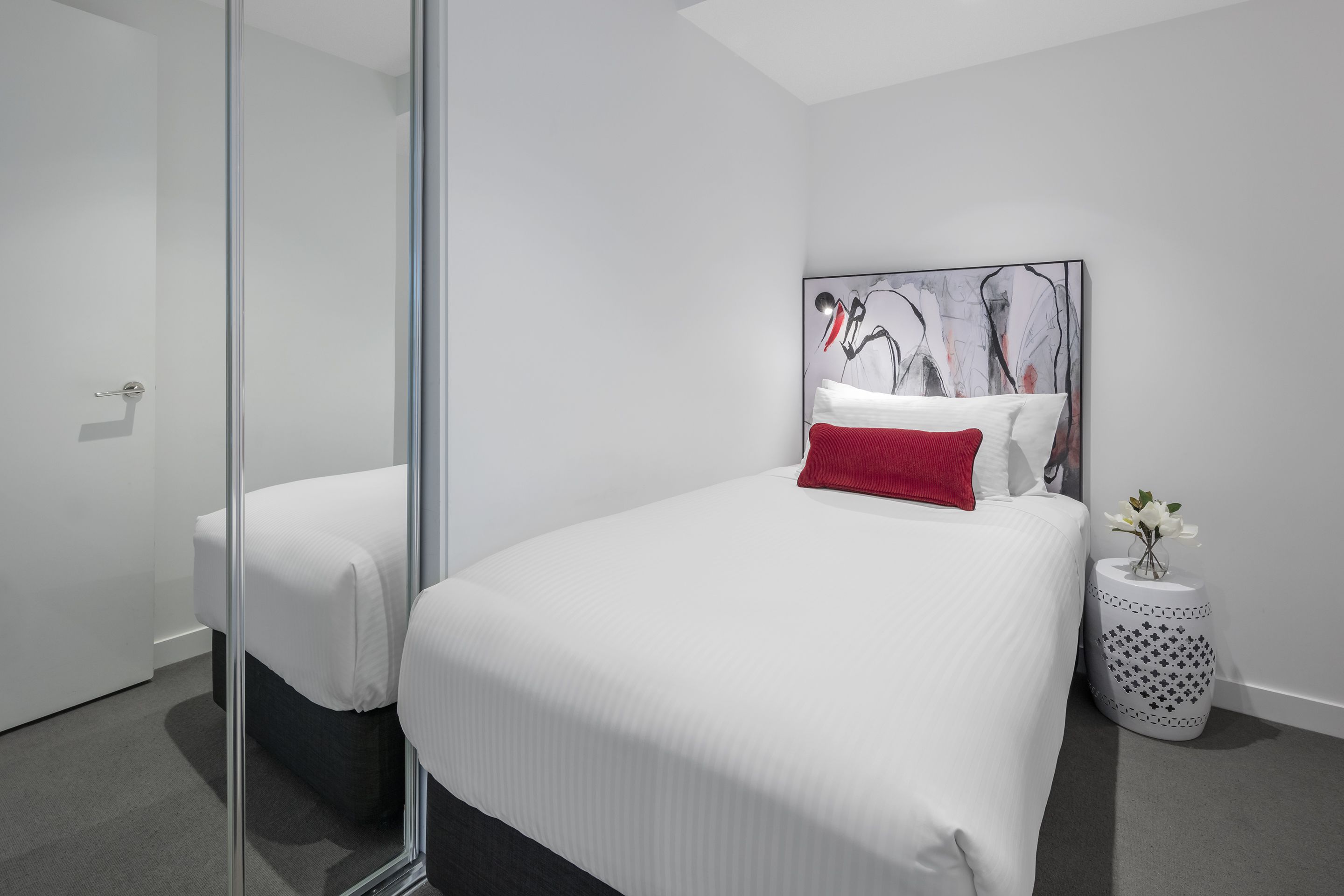 A modern hotel room with a large bed, a glass partition, a small side table with a vase, and a large abstract painting on the wall.