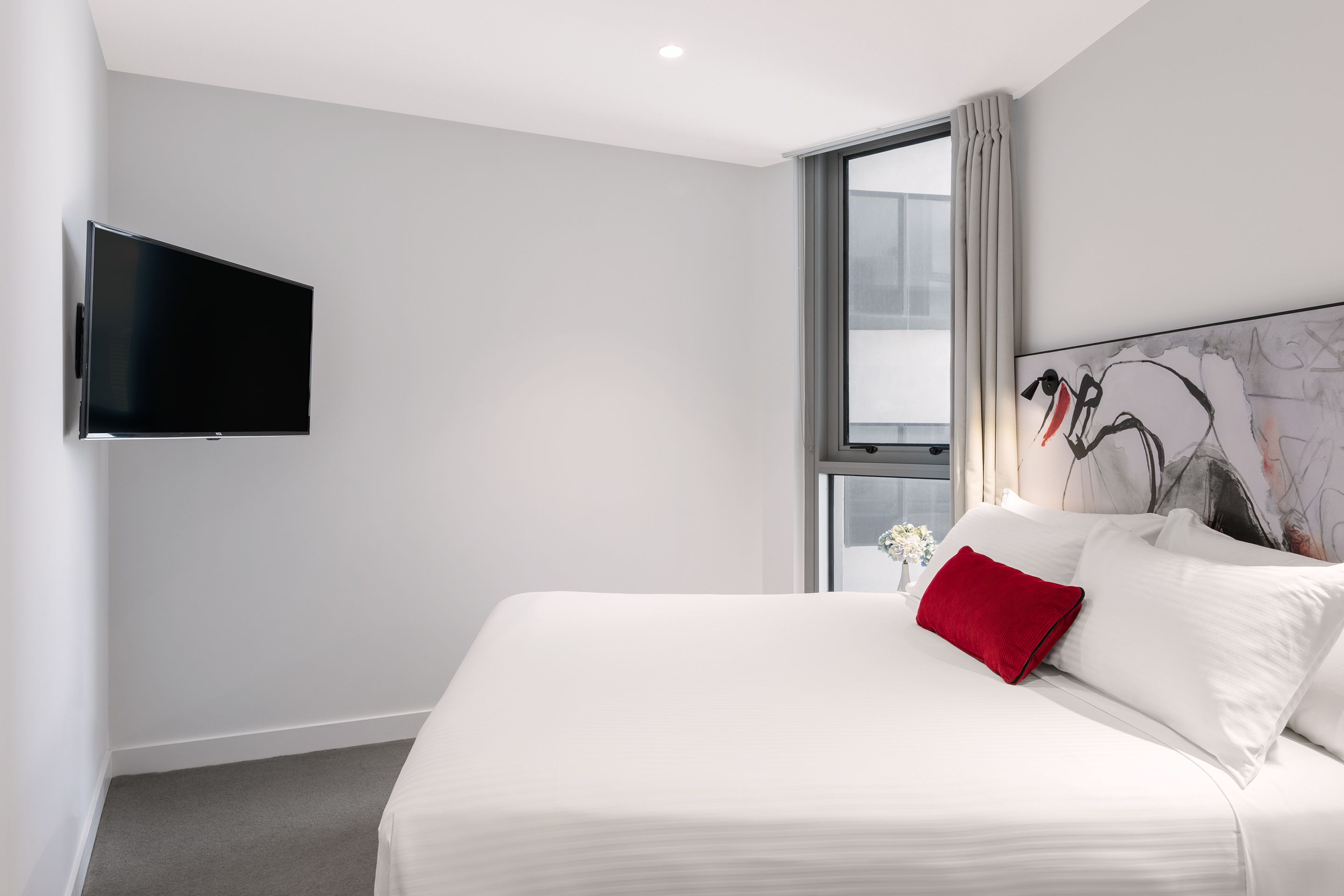 A modern hotel room with a large bed, a flat-screen TV mounted on the wall, and a window with curtains.