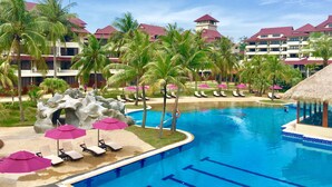 A resort pool area with lounge chairs and umbrellas, surrounded by palm trees and resort buildings. - Sand & Sandals Desaru Beach Resort & Spa (Bandar Penawar)