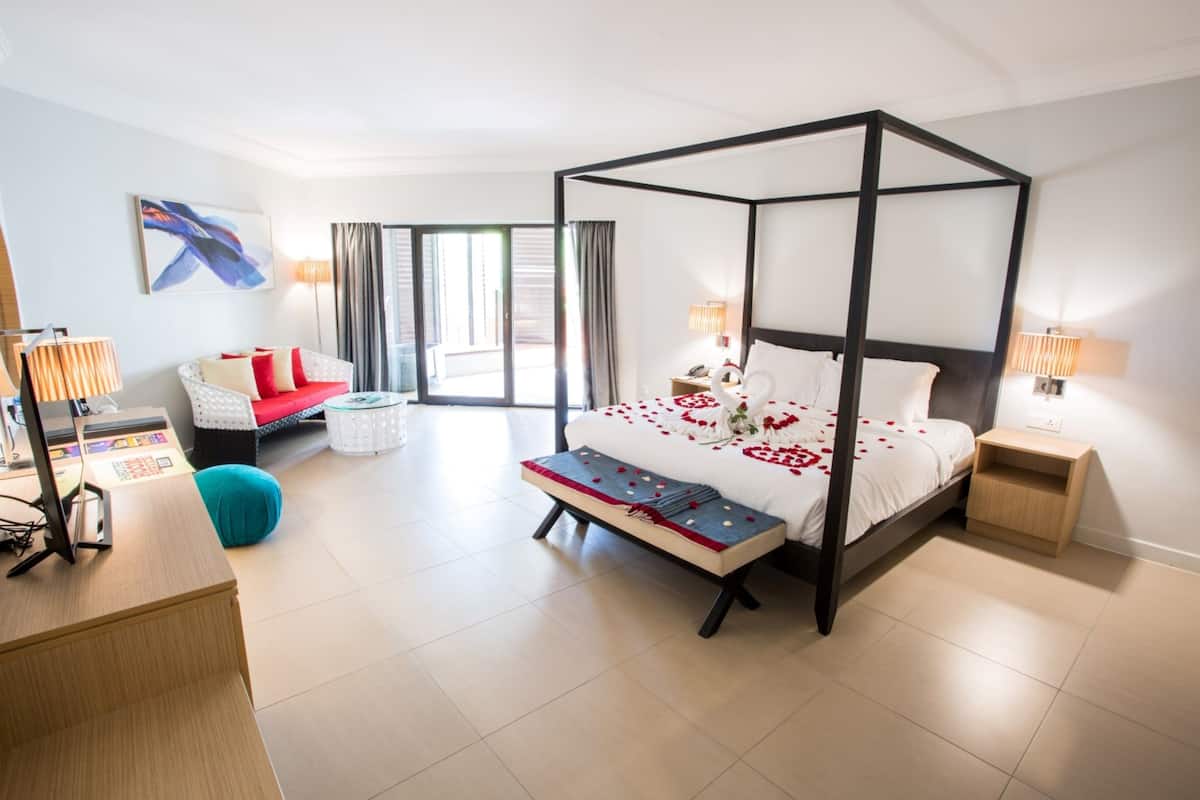 A modern hotel room with a four-poster bed, a sofa, a desk, and a large window.