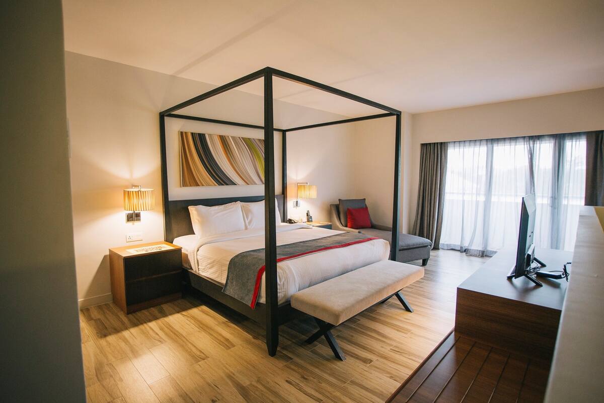 A modern hotel room with a four-poster bed, a desk with a computer, a chair, and a bench.