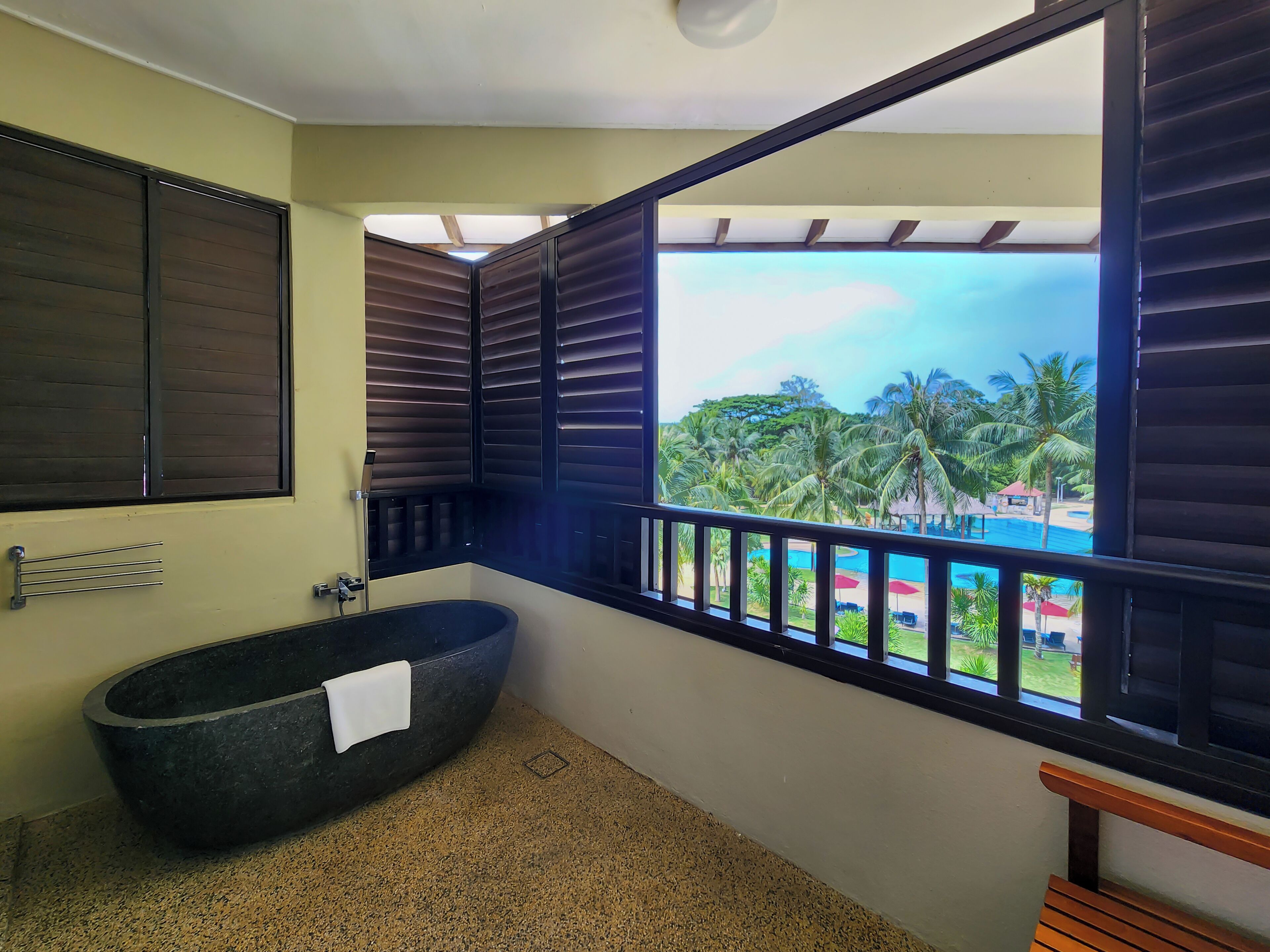 A bathroom with a freestanding bathtub, a towel rack, and a view of a pool and palm trees.