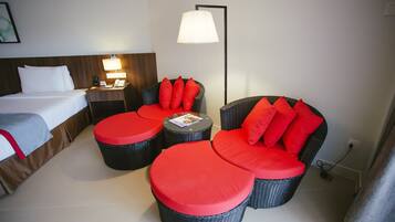 Habitación de hotel con cama, escritorio con lámpara, silla con cojines rojos y lámpara de pie.