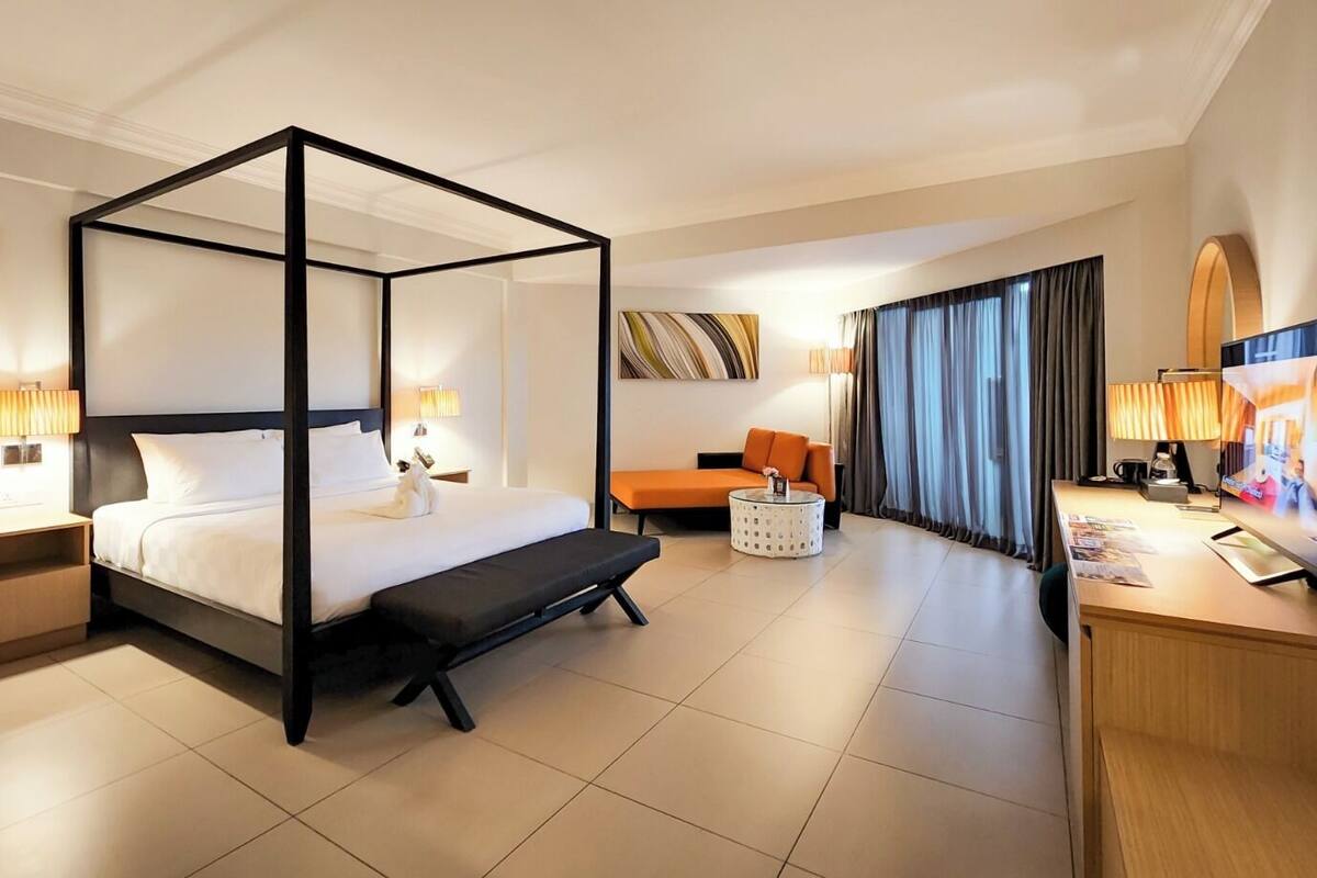 A modern hotel room with a four-poster bed, a sofa, a coffee table, and a TV.