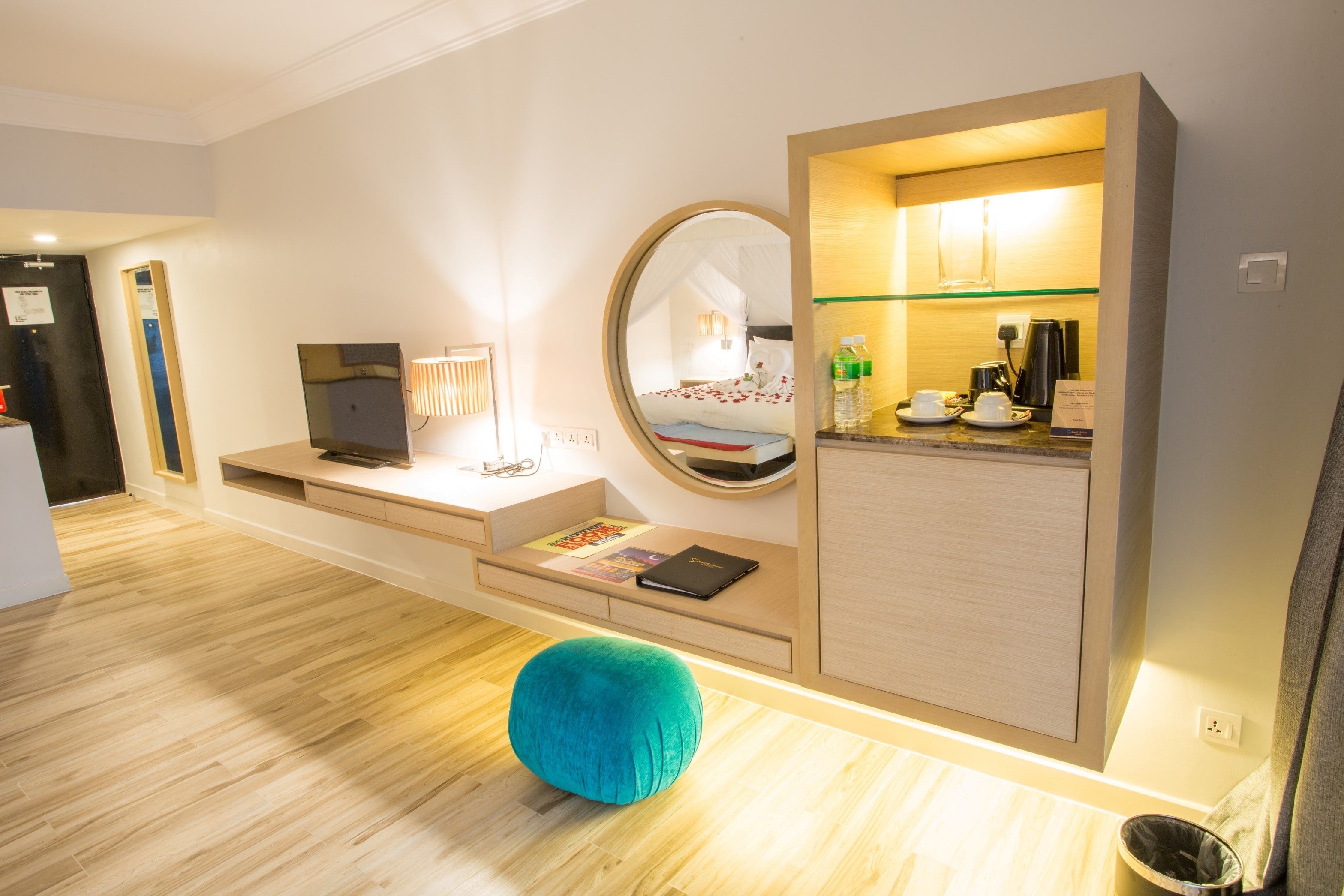 A modern hotel room with a wooden floor, a teal ottoman, a desk with a laptop, a mirror, and a kitchenette area.