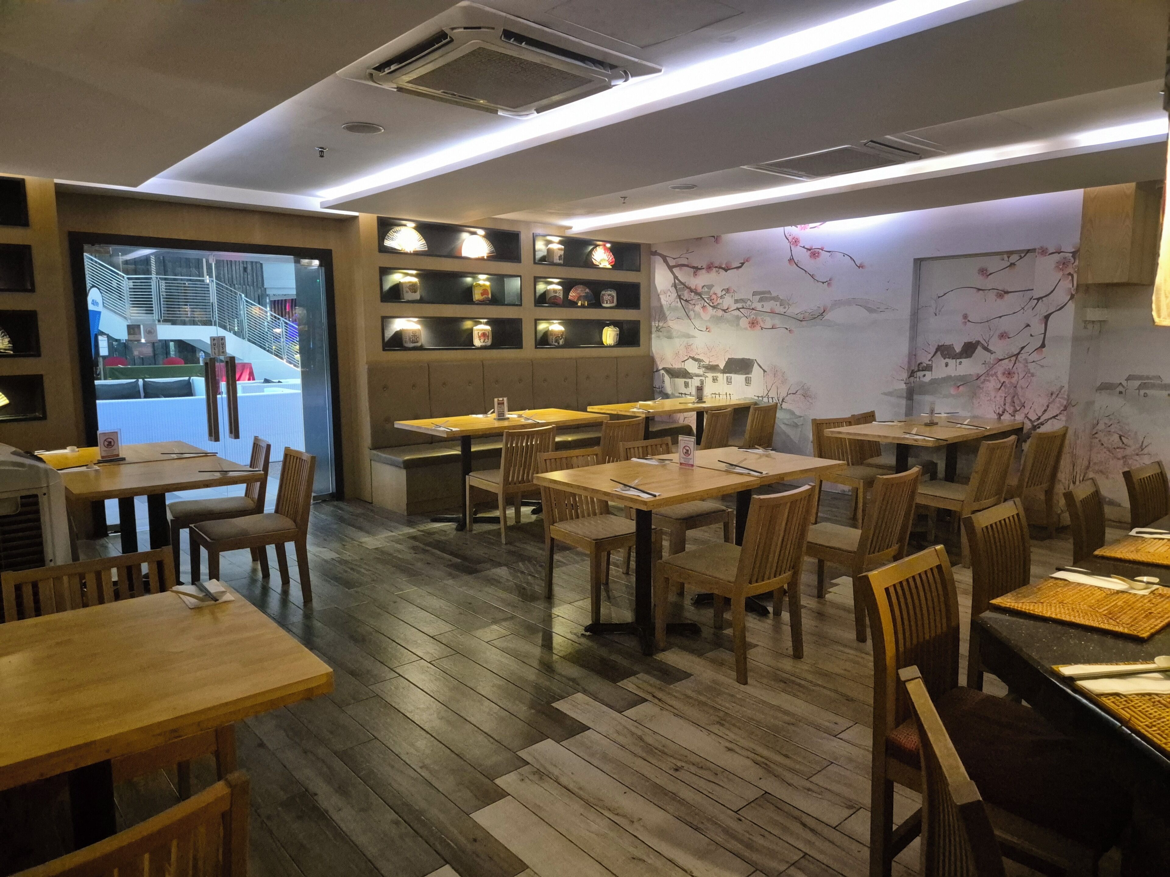 A restaurant interior with wooden tables and chairs, a wall-mounted menu board, and a view of the outside.