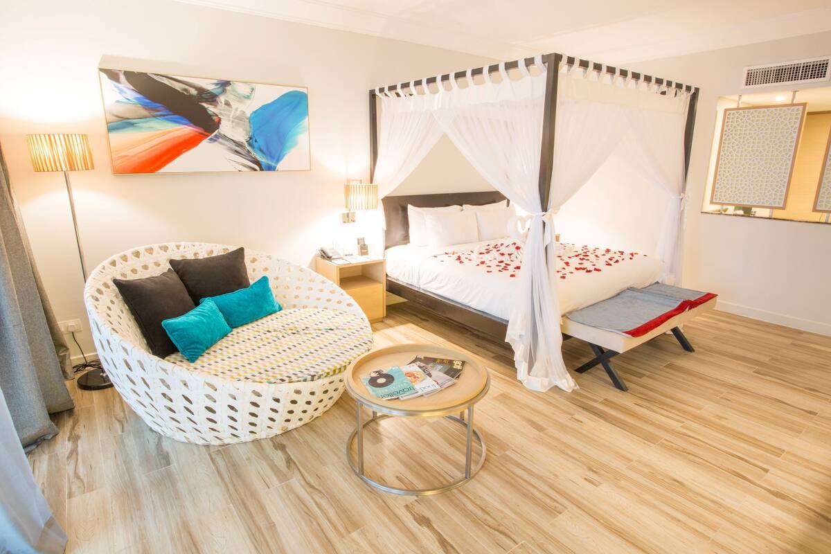 A bedroom with a four-poster bed, a round wicker chair, a small round table, and a wall-mounted lamp.
