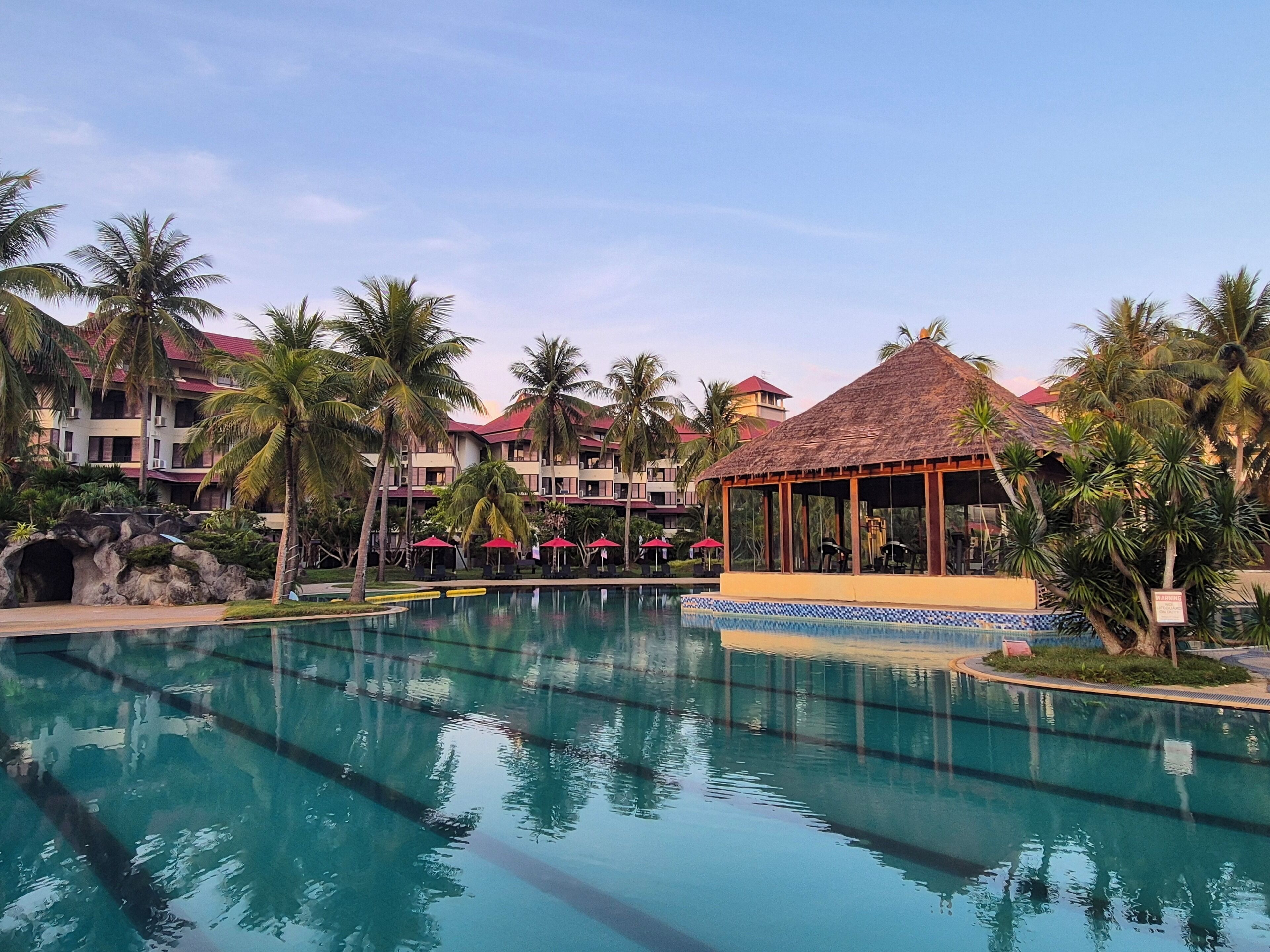 A resort with a swimming pool, palm trees, and a thatched-roof gazebo.