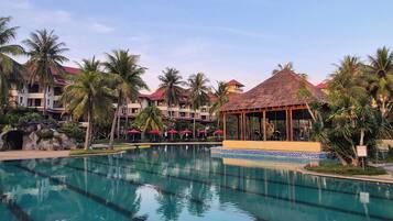 A resort with a swimming pool, palm trees, and a thatched-roof gazebo.