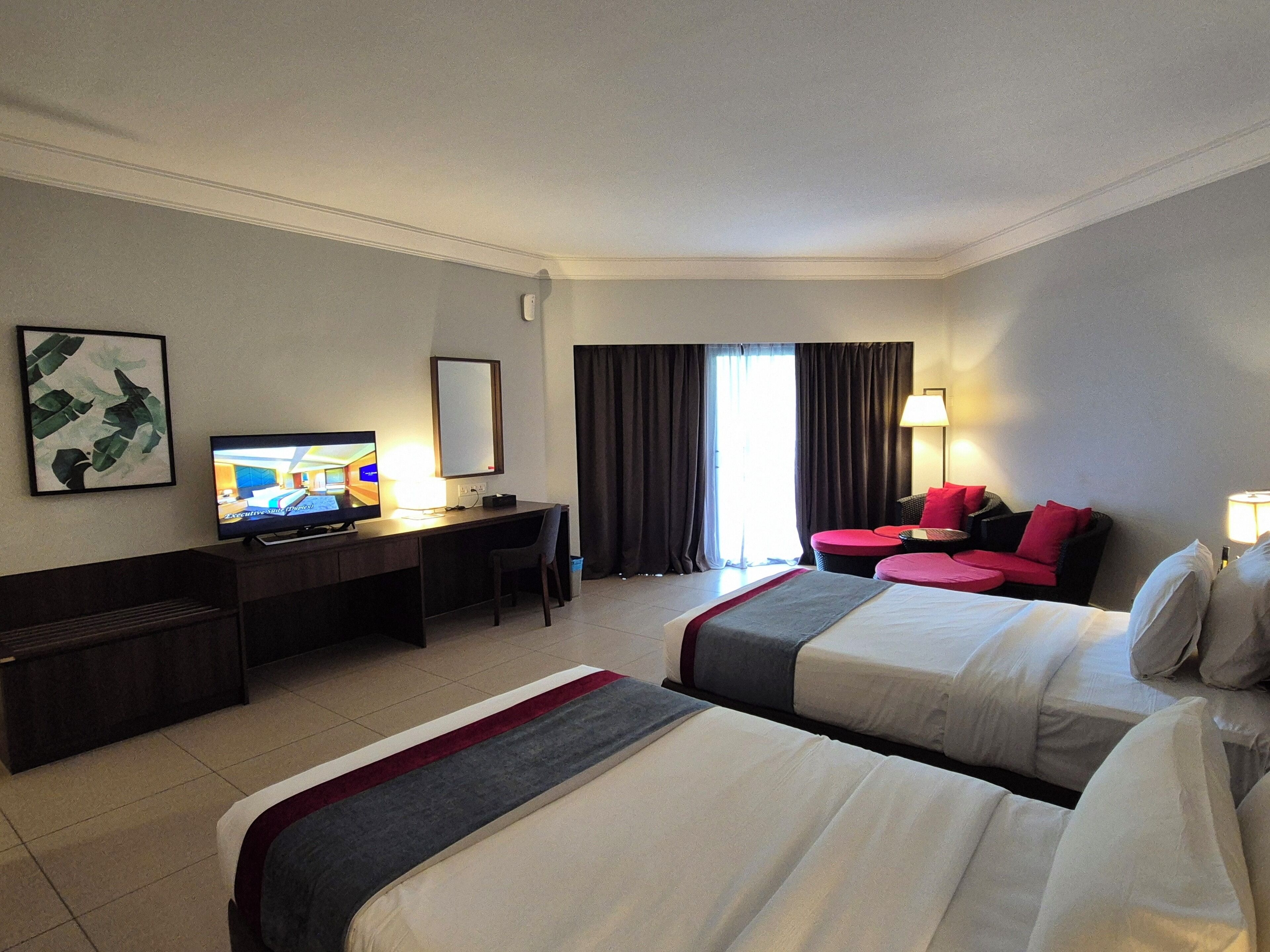 A hotel room with two beds, a desk with a television, a chair, a sofa, and a window with curtains.