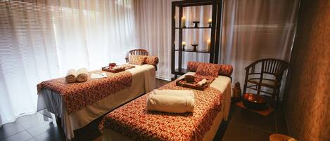 Un cuarto de spa con dos camas, una silla de madera, un estante con velas y un ventanal con cortinas translúcidas.