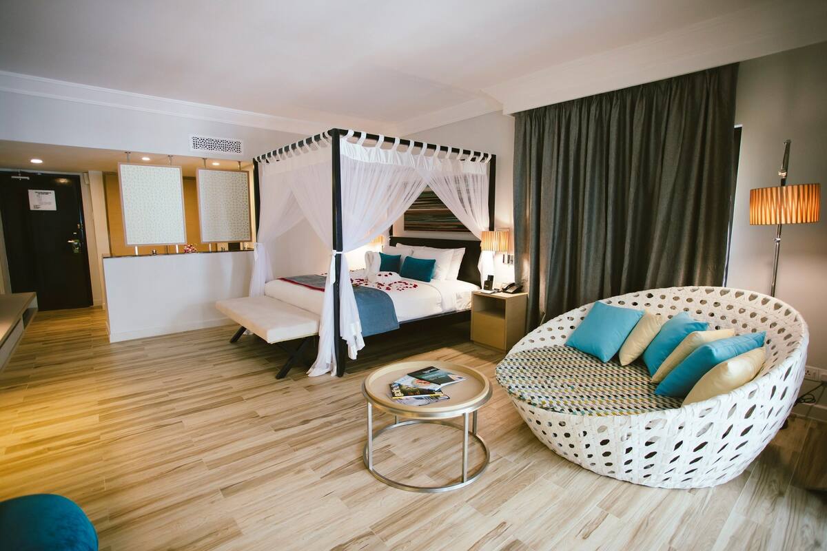 A bedroom with a four-poster bed, a round wicker sofa, a small round table, and a nightstand with a lamp.