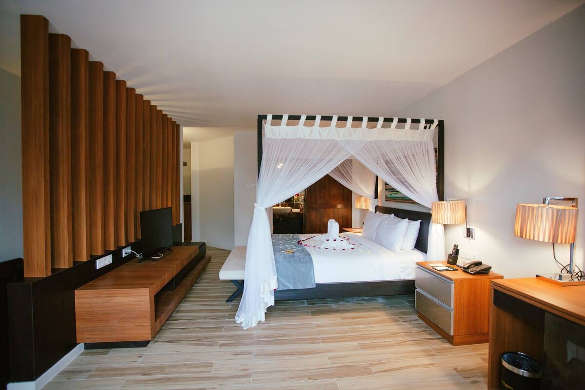 A modern hotel room with a large bed, a bedside table, a TV, and a wooden bench.