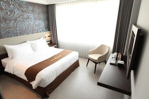 Habitación de hotel con cama, una silla, escritorio con televisor y ventana con cortinas.