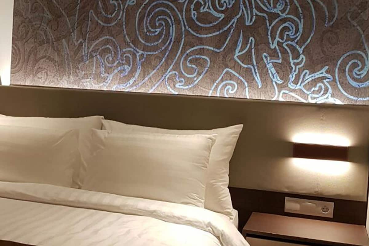A neatly made bed with white linens and a brown throw blanket, a bedside table with a lamp, and a wall with a decorative pattern.