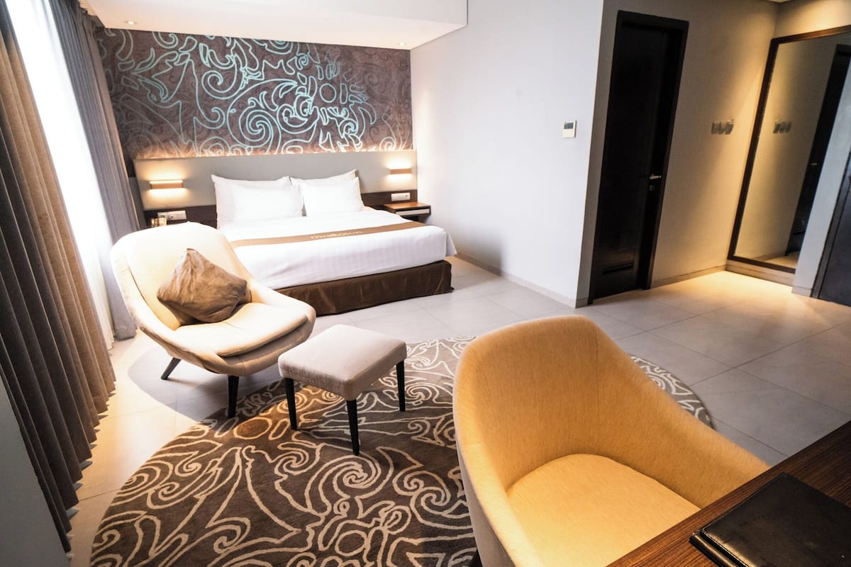 A modern hotel room with a large bed, a sitting area with a chair and ottoman, a patterned rug, and a large mirror.