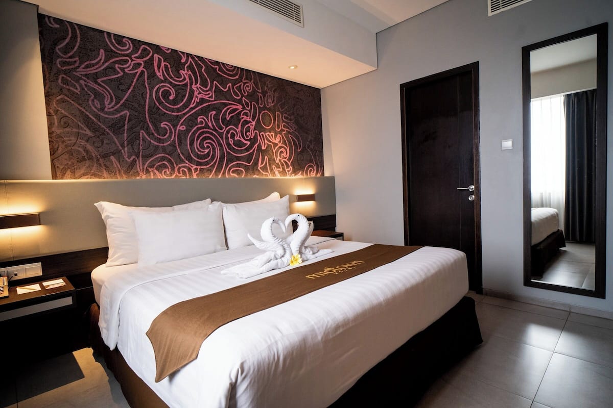 A hotel room with a large bed, a headboard with a decorative panel, a bedside table with a lamp, and a large mirror.