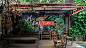 Et rustikt udendørs siddeområde med træbænke og borde, omgivet af frodigt grønt og et muralmaleri med jungletema.