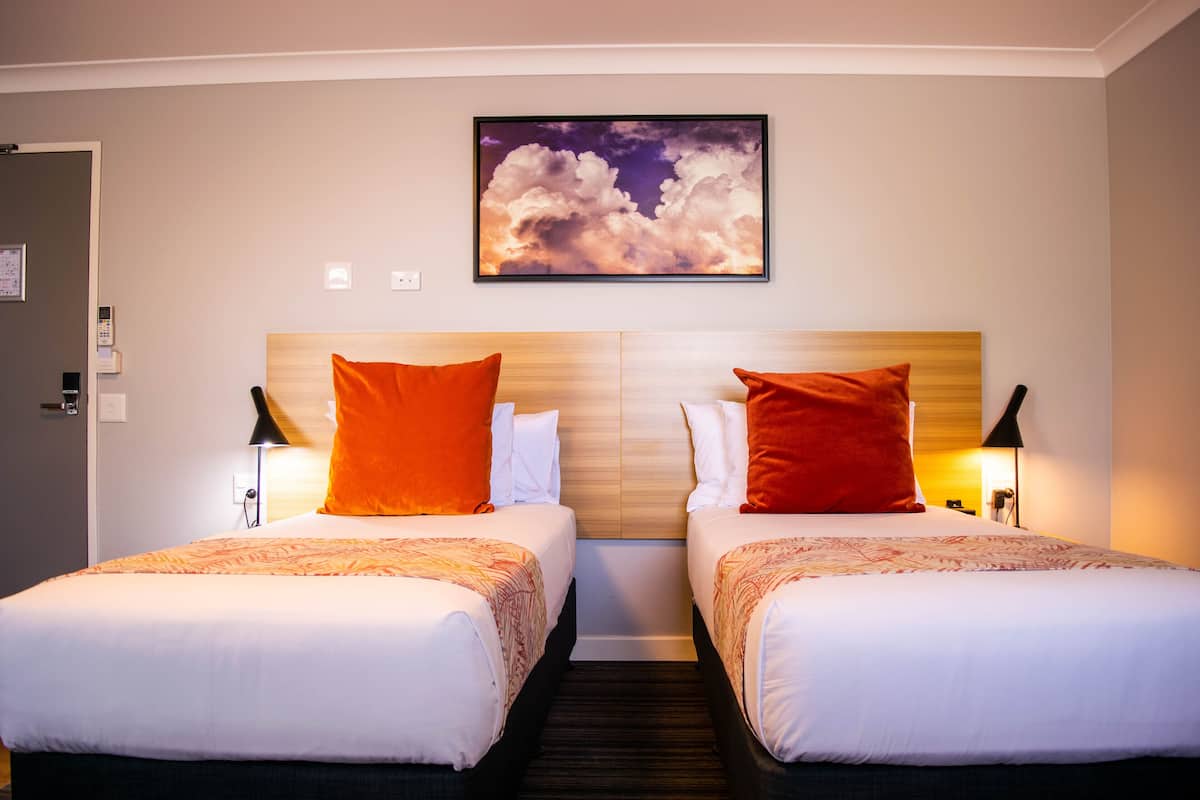 Une chambre d’hôtel avec deux lits, chacun doté d’une tête de lit en bois, de draps blancs et d’oreillers orange. Un tableau encadré représentant des nuages est accroché au mur, au-dessus des lits.
