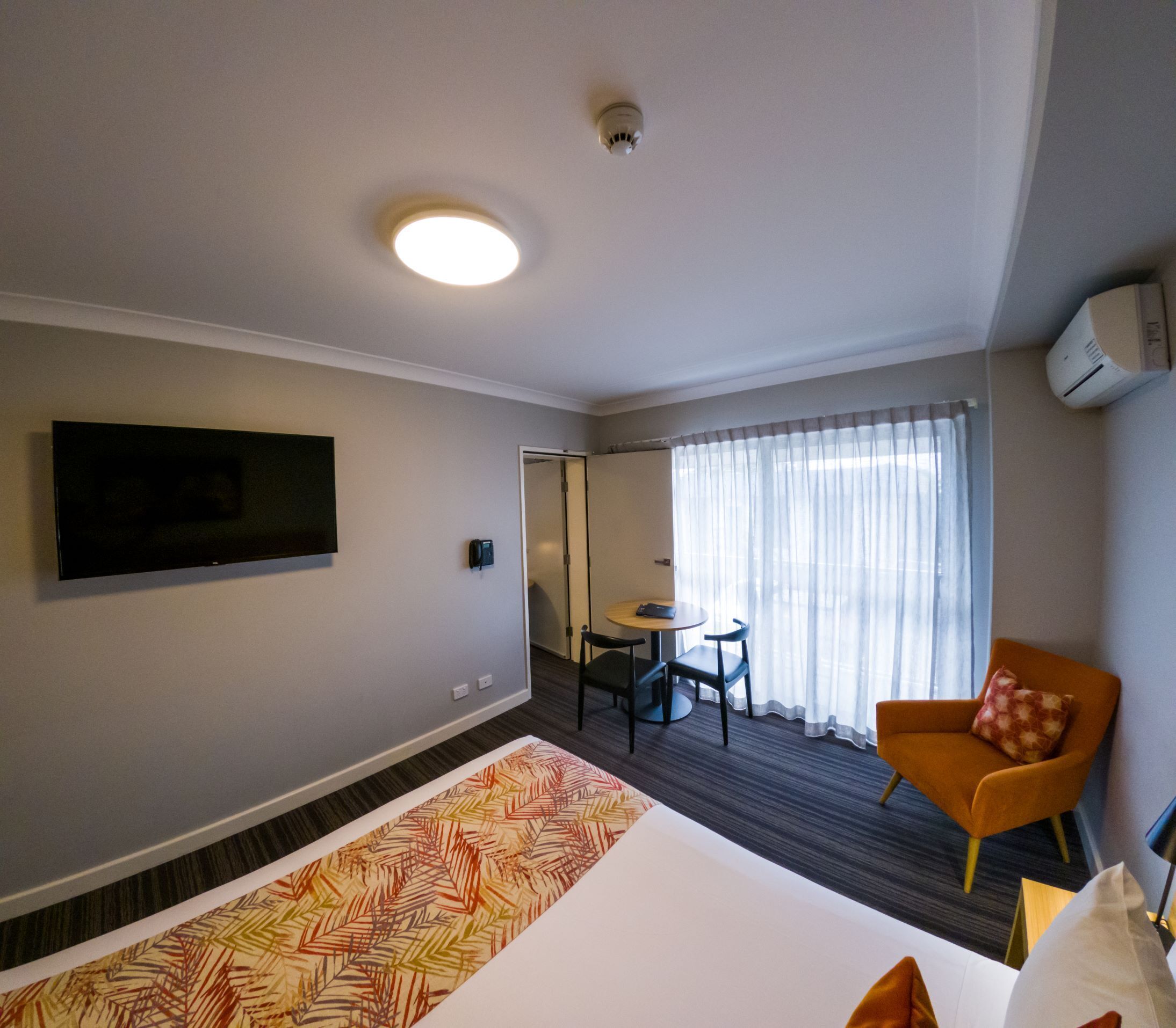 Une chambre d’hôtel avec un grand lit, un téléviseur fixé au mur, une petite table avec des chaises et une fenêtre avec des rideaux.