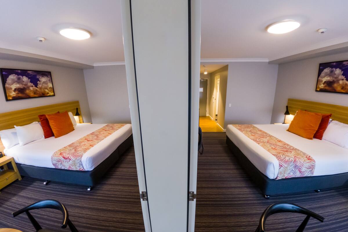 Une chambre d’hôtel aux murs recouverts de miroirs, avec deux lits, une tête de lit en bois, une table de chevet, une chaise et un couloir menant à une autre pièce.