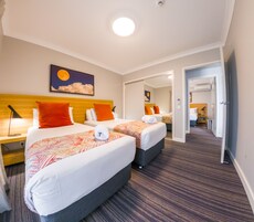 Quarto de hotel com duas camas, uma mesa de cabeceira com abajur, um quadro com lua e nuvens, e piso coberto por carpete.