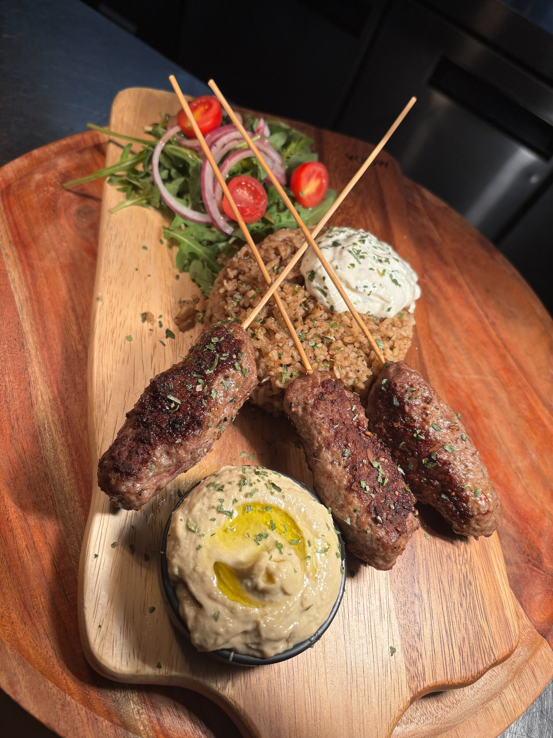 A wooden board with kebabs, a salad, and a bowl of hummus.