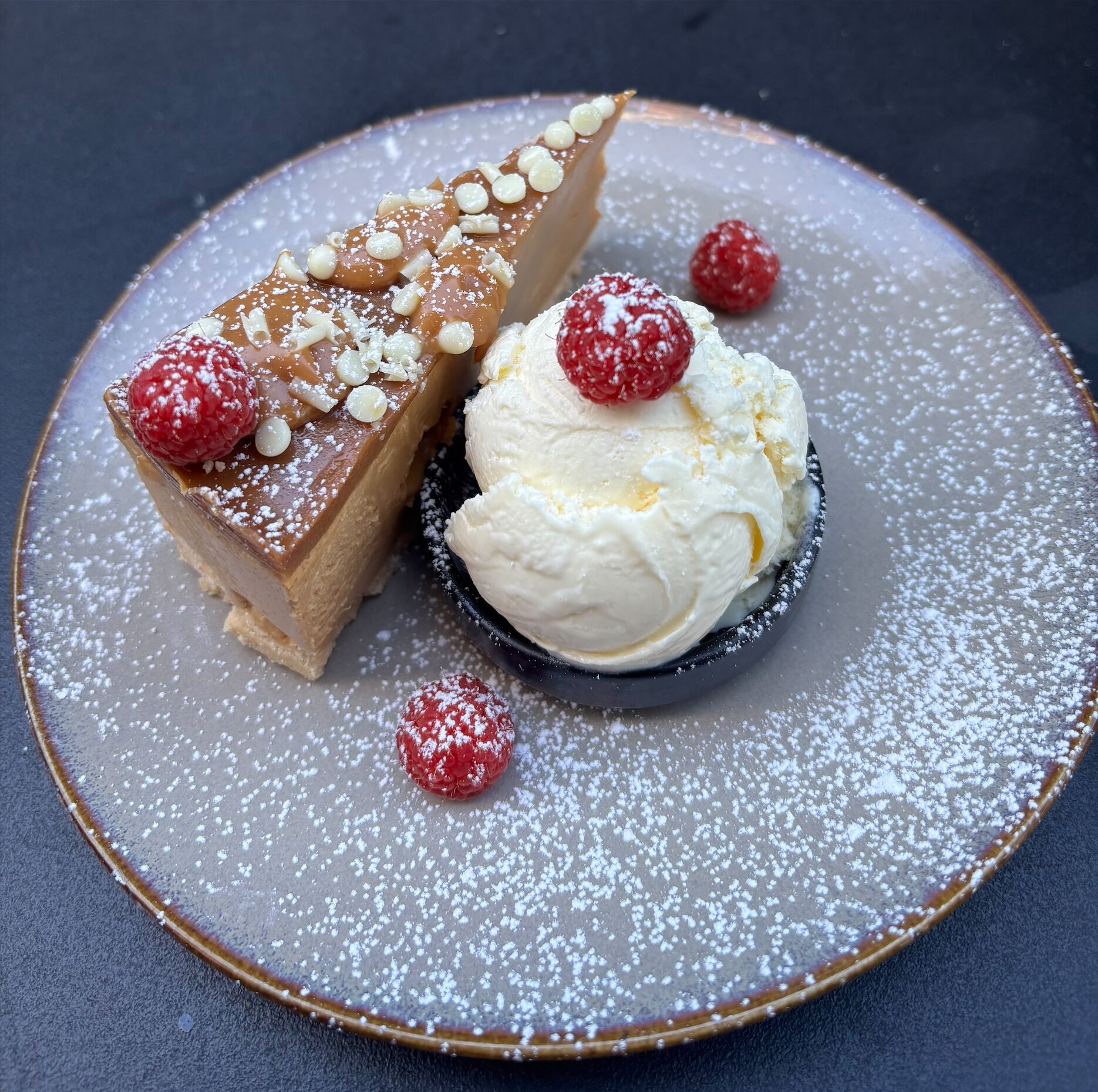 Un plato de postre con una porción de pastel cubierto de nueces y azúcar glas, una bola de helado de vainilla y una frambuesa.