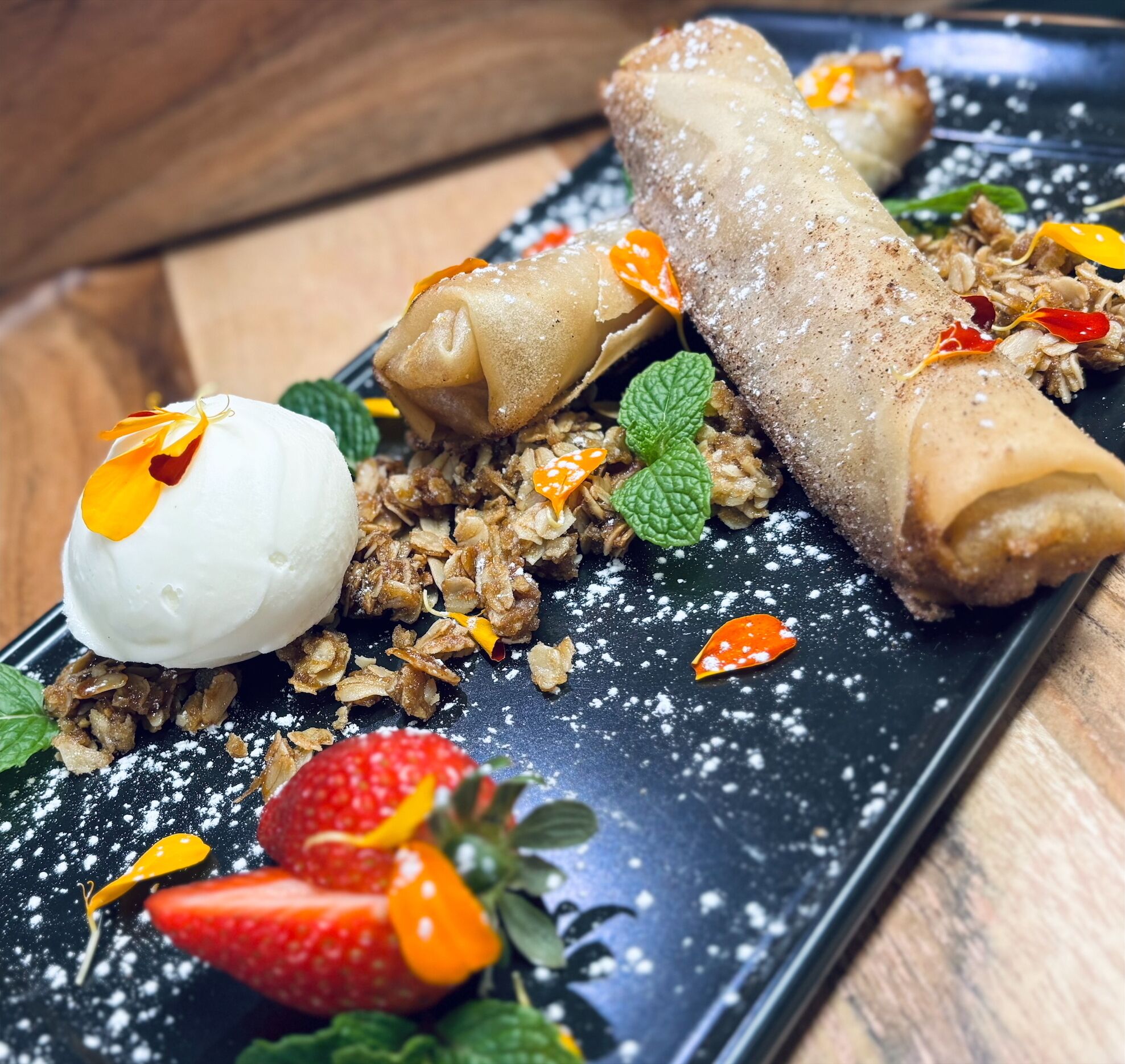 Une assiette de dessert avec des pâtisseries roulées, une boule de glace, du granola, des fraises et des feuilles de menthe.