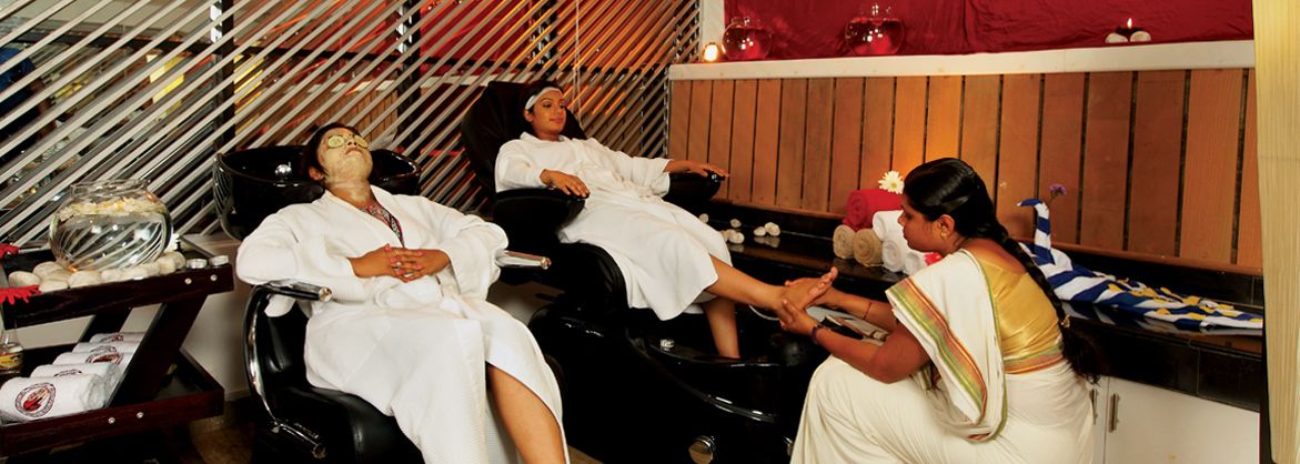A traditional spa setting with two clients in black chairs, a masseuse in traditional attire, and a decorative fish bowl.