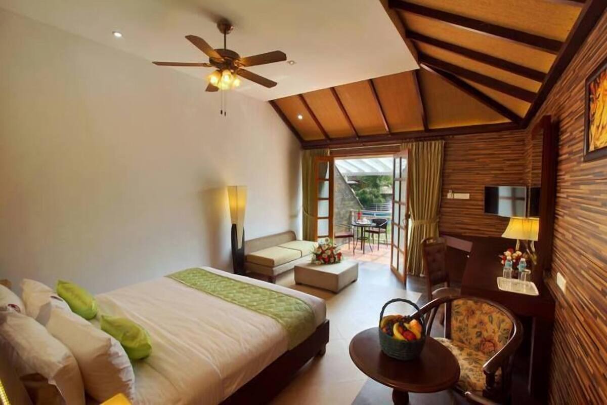 Quarto de hotel com cama, escrivaninha, cadeira, ventoinha no teto e vista para o exterior.