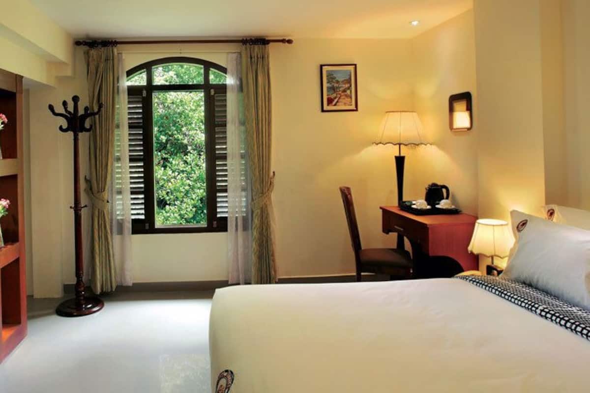 Quarto de hotel com cama, televisão presa a um painel de madeira, escrivaninha com abajur e janela com cortinas.
